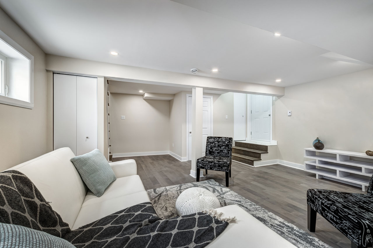 A clean, contemporary finished basement featuring a white sofa, patterned chairs, wooden flooring, recessed lighting, and a staircase leading to the upper level.