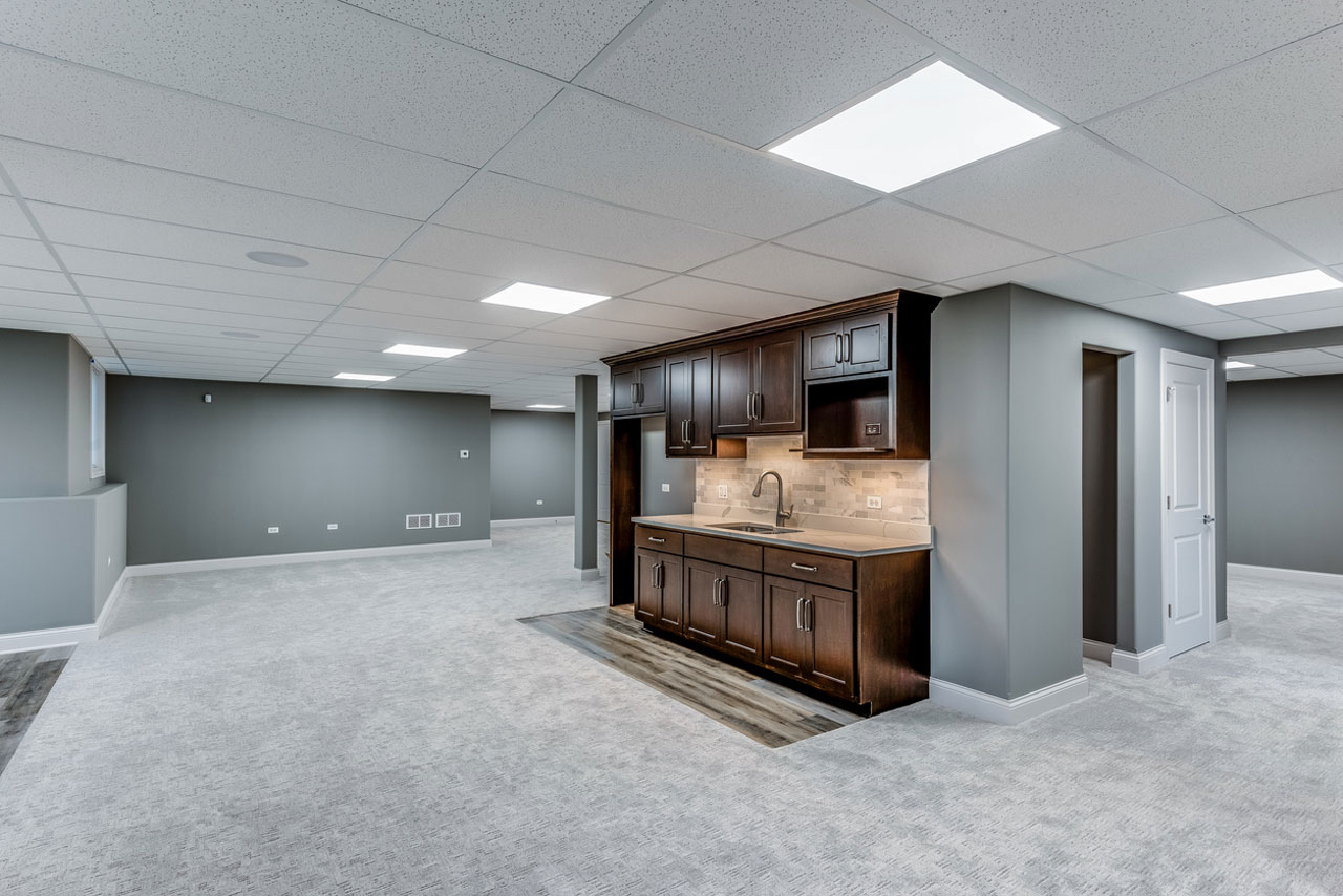 A clean, finished basement with light-colored walls, hardwood-style flooring, a central staircase with black railings, and a cozy seating area, ideal for family gatherings.