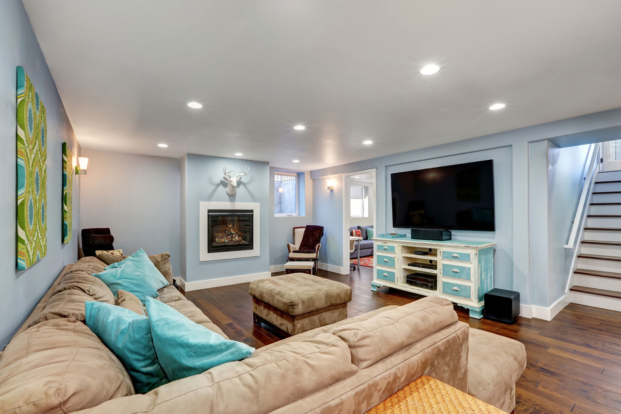A warm and inviting basement family room featuring a fireplace, large TV on a blue cabinet, comfortable couches with teal pillows, and a staircase leading up, perfect for relaxation and entertainment.
