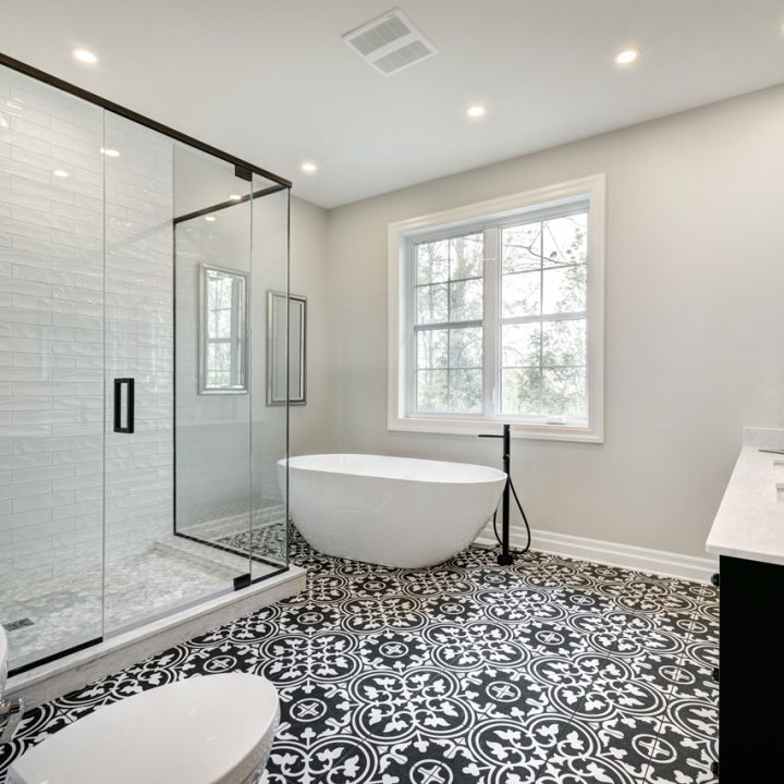 A stylish bathroom featuring a freestanding soaking tub, glass-enclosed shower with white subway tile, black and white patterned floor tiles, dual sinks, and a large window allowing natural light.