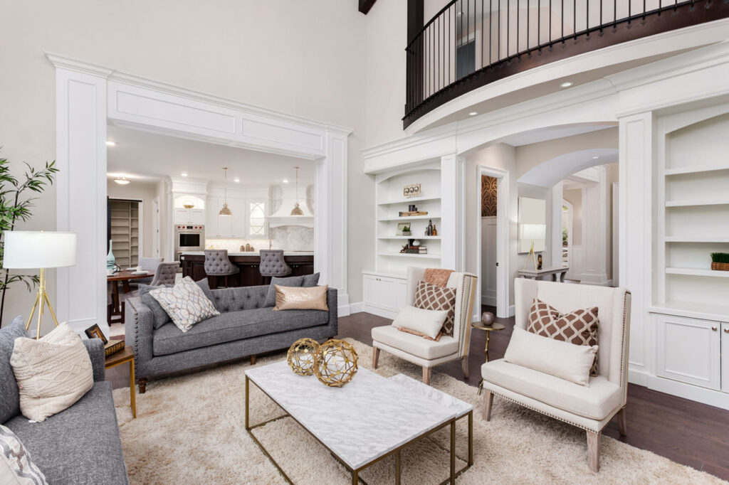 A spacious, well-lit living room with high ceilings, a balcony above, white trim, built-in shelves, a gray sofa, and a marble-top coffee table, showcasing refined design.