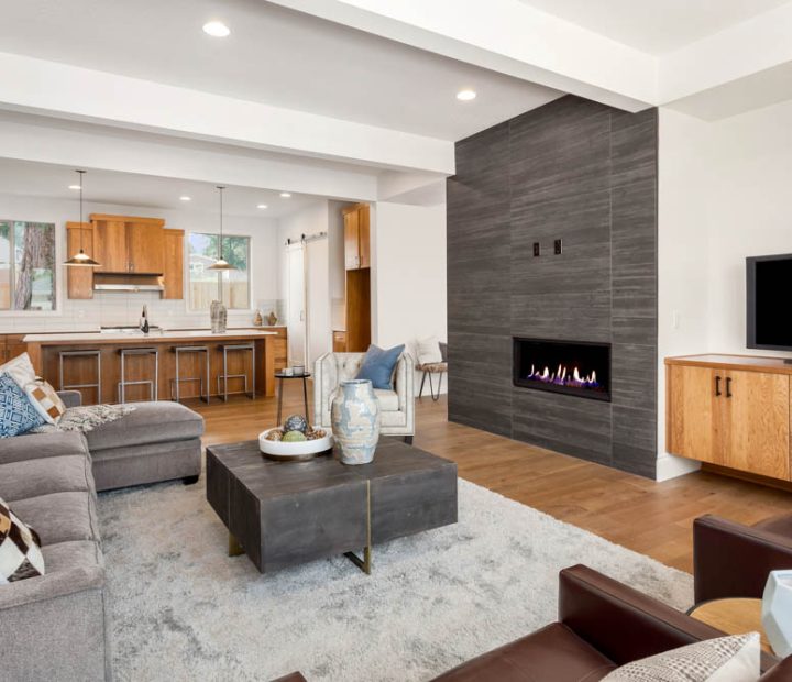 A stylish living area with a sleek linear fireplace, gray sofas, a dark wood coffee table, hardwood flooring, and an open view into a warm-toned kitchen.