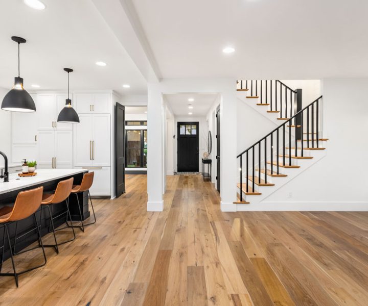 A bright, airy home interior with a black island kitchen, white cabinetry, leather bar stools, hardwood floors, and a floating staircase with black metal railings.