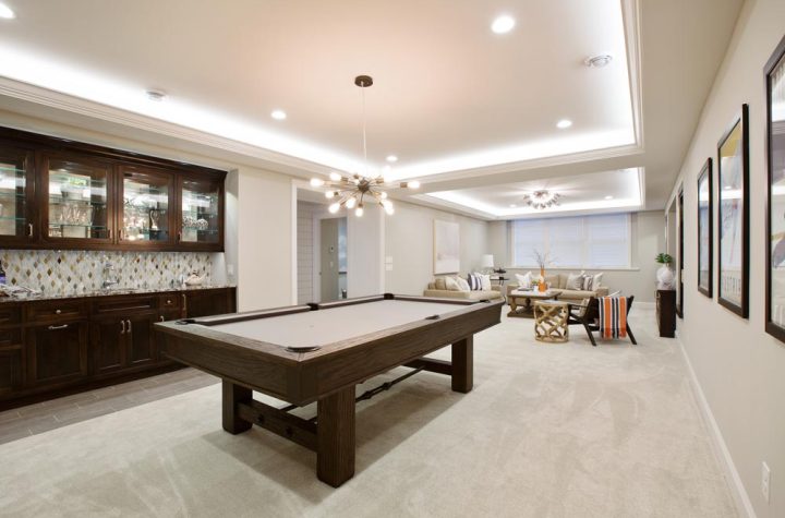 A spacious, finished basement featuring a wooden pool table, built-in bar with dark cabinets and mosaic backsplash, comfortable seating, and modern lighting for entertainment.