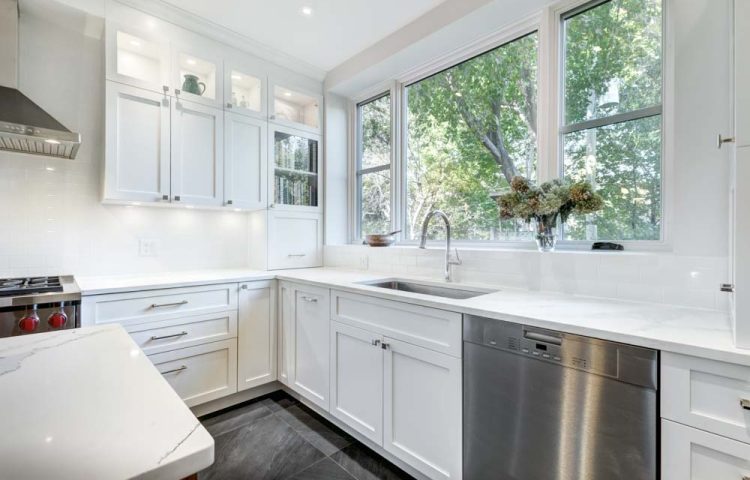 A spacious, airy kitchen with white cabinetry, quartz countertops, stainless steel appliances, large windows overlooking greenery, and a built-in sink with a modern faucet.