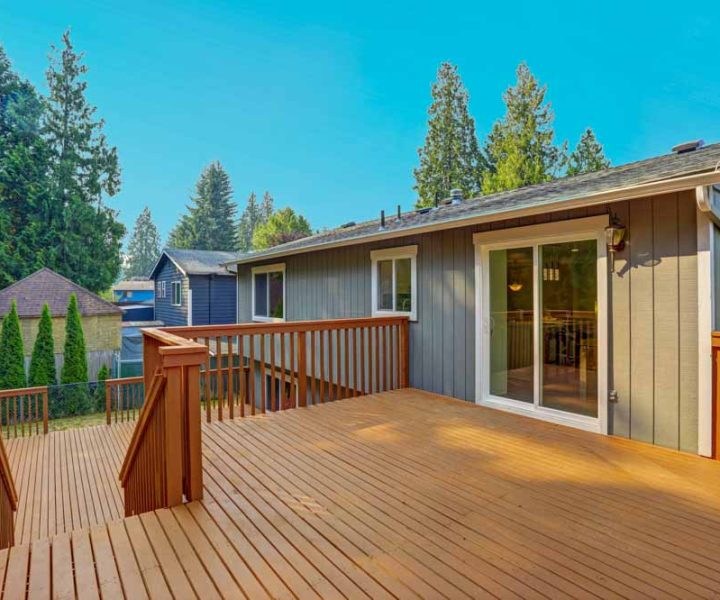 A large, well-lit wooden deck attached to a modern home with a sliding glass door, featuring warm-toned railings and a scenic view of trees and neighboring houses.